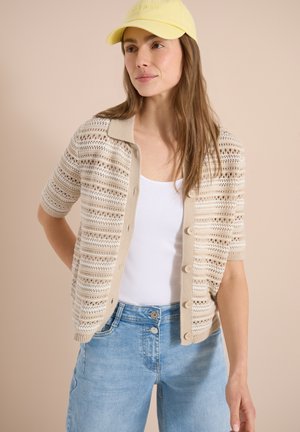 Kvinde iført gul kasket, beige åbenstrikket kortærmet cardigan, hvid tanktop og lyseblå højtaljede jeans mod en beige baggrund.