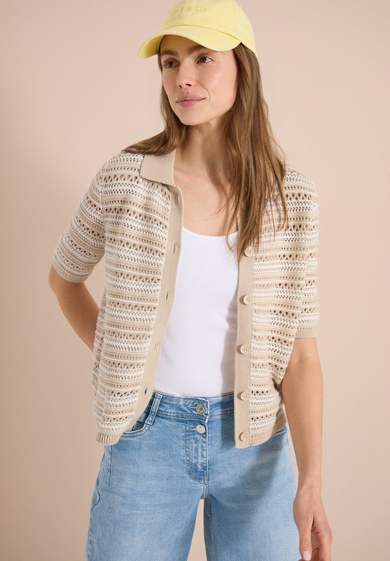 Femme portant une casquette jaune, un cardigan beige en maille ajourée à manches courtes, un débardeur blanc et un jean taille haute bleu clair sur fond beige.