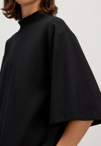 Zwarte oversized top met een hoge nek, korte wijde mouwen en een gladde textuur, met een minimalistisch ontwerp en subtiele stiksels.