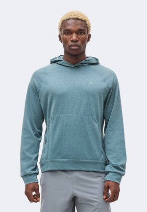 Modèle masculin aux cheveux blonds courts portant un sweat à capuche bleu sarcelle à manches longues et un short gris clair, debout devant un fond uni.