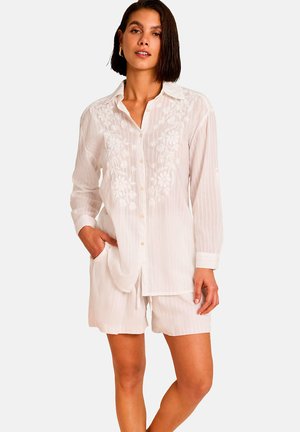 Femme portant une chemise blanche à manches longues avec broderie florale et un short rayé assorti, debout avec une main dans la poche.