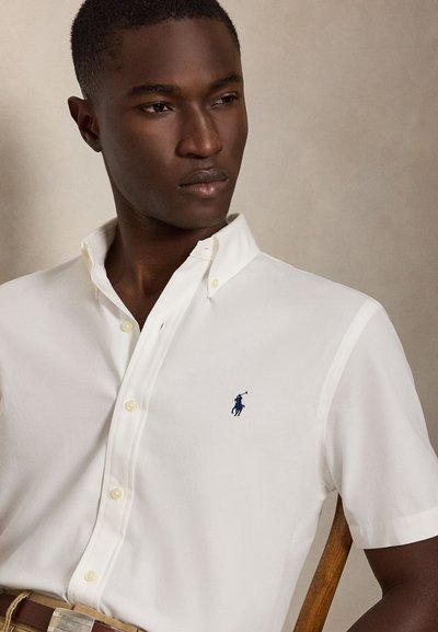 Mann iført en hvit kortermet skjorte med en liten marineblå polospiller-logo på brystet, sittende på en trestol.