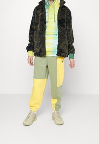 Fleecejacke in Dunkelgrün mit schwarzer Textur, über einem gelb-grünen Hoodie mit Batikmuster getragen. Jogginghose in Grün und Gelb mit seitlichen Einsätzen.