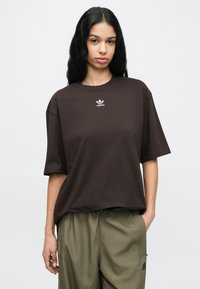 Γυναίκα με μακριά σκούρα μαλλιά που φοράει καφέ μπλουζάκι Adidas και ελιά πράσινο παντελόνι Adidas, στέκεται με το ένα χέρι στην τσέπη.