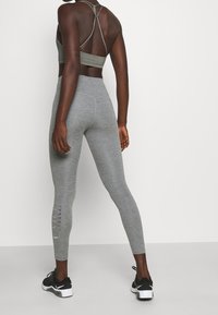 Šedá sportovní podprsenka s křížovými zadními popruhy spárovaná se šedými legínami s bočním výřezem a černými sportovními botami.