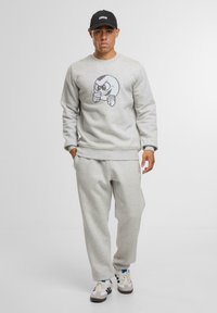 Felpa grigia chiara con una grafica di personaggio dei cartoni animati, abbinata a pantaloni grigi coordinati. Comprende un berretto nero con la scritta "UNFAIR".