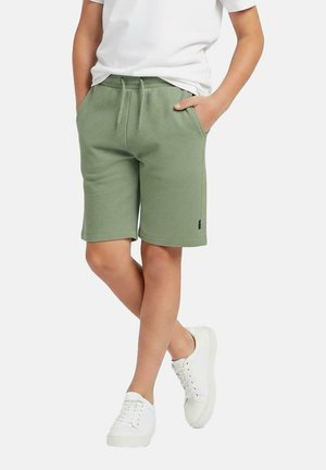 Kind draagt lichtgroene short met trekkoord, wit T-shirt en witte sneakers, staat met gekruiste benen en handen in de zakken.