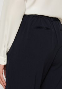Gros plan d'une personne portant un pantalon bleu marine avec des poches arrière et une chemise blanche à manches longues rentrée avec des poignets boutonnés.