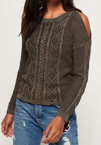 Svetr z pletené olivově zelené vlny s otevřeným designem ramen, zdobený texturovaným kabelovým vzorem a žebrovanými manžetami. Kombinovaný s roztrhanými džíny.