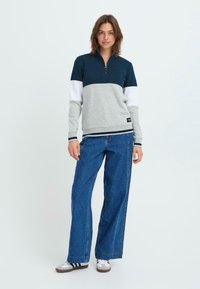 Felpa con zip a blocchi di colore grigia, blu navale e bianca, con maniche lunghe e orlo a coste, abbinata a jeans a gamba larga blu e sneakers.