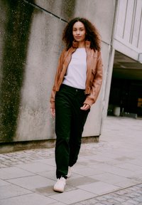 Woman with curly hair walking outdoors wearing a brown leather jacket, white shirt, black pants, and white sneakers by a concrete wall.