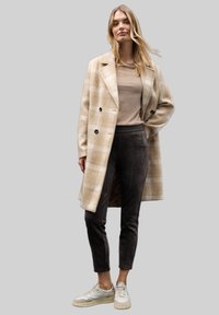 Beige rutigt ullblandat ytterplagg med dubbelknäppning, kombinerat med mörkgrå mockabyxor och krämfärgade sneakers. Slät textur och skräddarsydd passform.