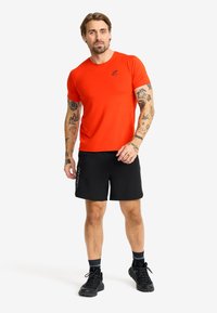 Herrträningsoutfit bestående av en figursydd orange t-shirt och svarta shorts. T-shirten har en diskret logotyp; shortsen är lätta med sidodetaljer.