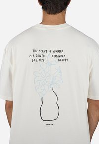 Hvid bomulds t-shirt med sort omrids af en vase med blå blomster. Teksten ovenfor i fed sort skrifttype med et citat om sommerens skønhed.