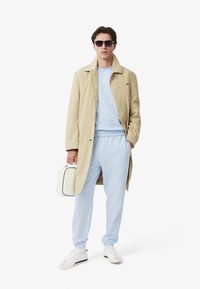 Bež trench coat preko svetlo modre dukserice in ujemajočih se trenirk. Bela torba z črnimi akcenti. Model nosi sončna očala in bele superge.