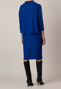 Pull bleu en maille avec manches trois-quarts et texture côtelée, associé à une jupe crayon bleue avec une fente arrière, complété par des bottes hautes noires jusqu'au genou.