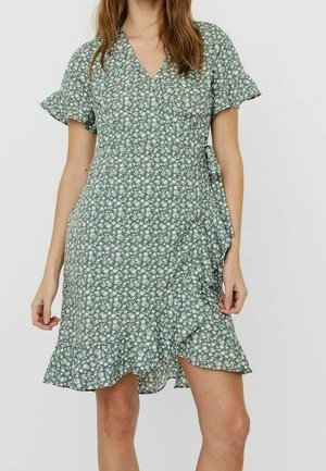 Woman wearing a green wrap dress with white floral print, short ruffle sleeves, and a ruffled hem, standing against a plain background.