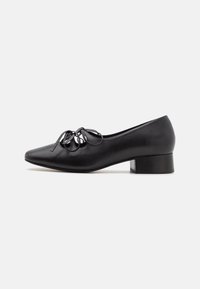 Zapato de cuero negro con tacón, de punta afilada, que presenta un detalle decorativo de lazo en la parte superior y un tacón bajo y cuadrado. Textura suave.