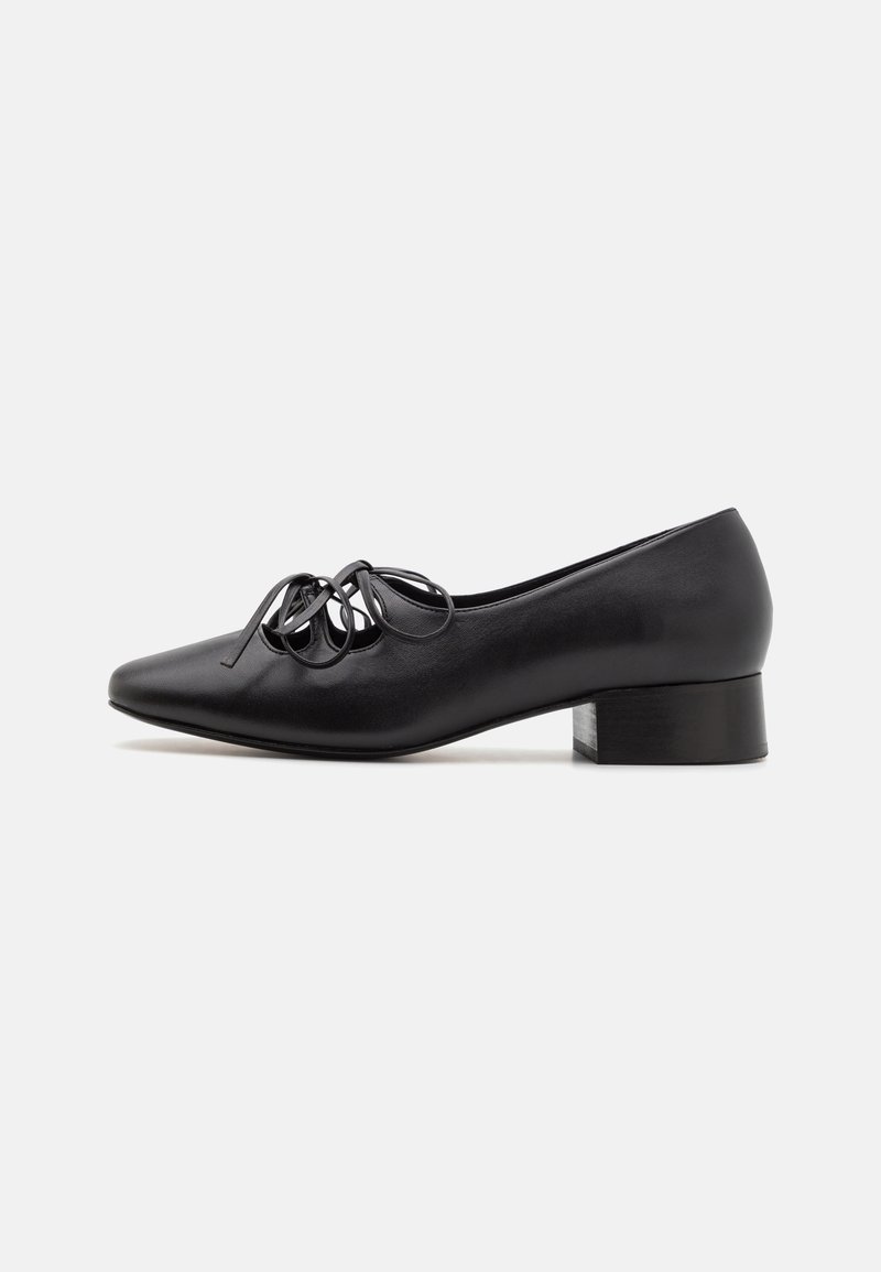Zapato de cuero negro con tacón, de punta afilada, que presenta un detalle decorativo de lazo en la parte superior y un tacón bajo y cuadrado. Textura suave.