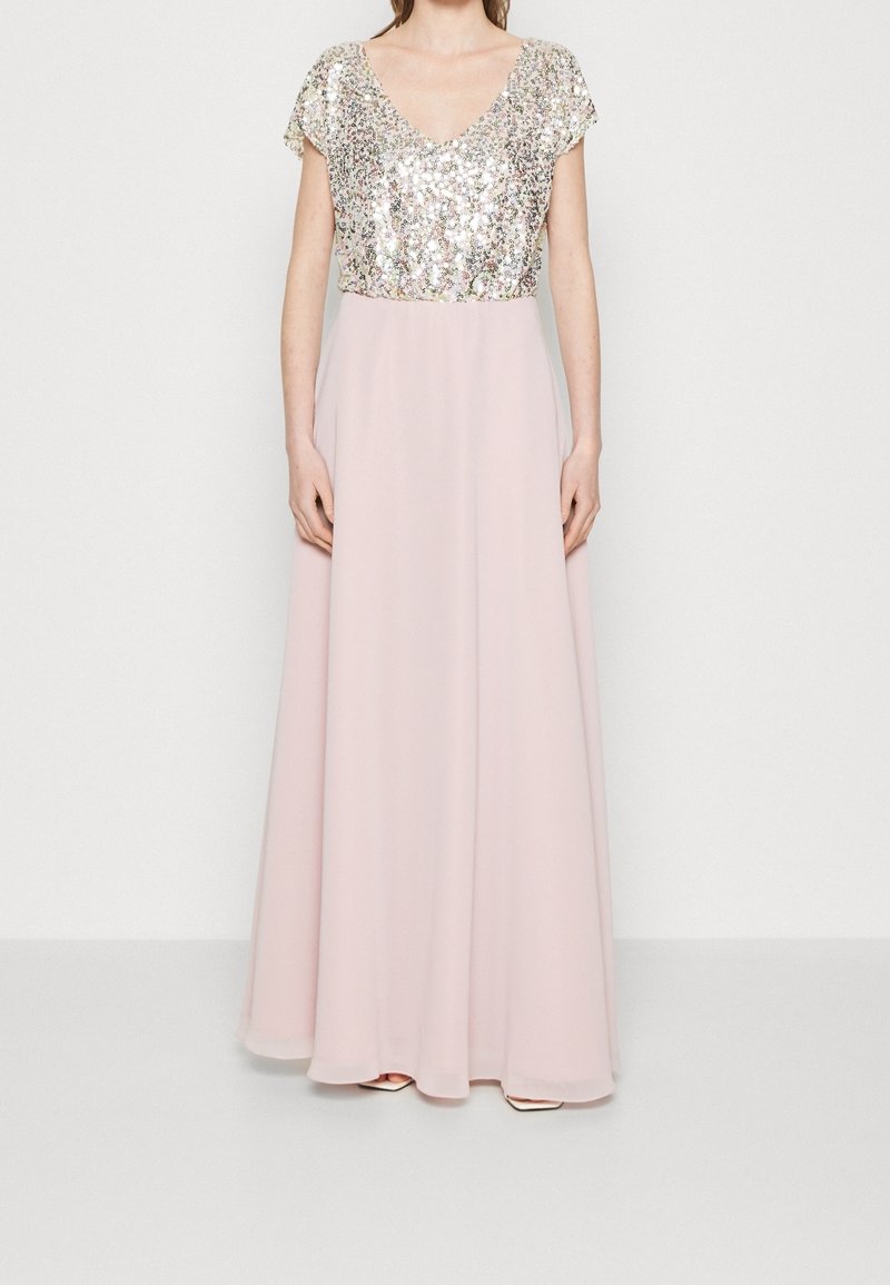 Femme portant une robe longue rose pâle avec un haut à sequins argentés à manches courtes et un décolleté en V, debout devant un fond uni.