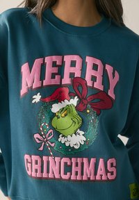 Teal színű pulóver, amelyen a Grincs egy ünnepi koszorúban látható. A felirat: "MERRY GRINCHMAS" rózsaszín és fehér színben. Puha, laza anyag.