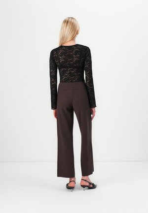 Γυναίκα με ξανθά μαλλιά, φοράει μαύρο δαντελένιο μακρυμάνικο τοπ, σκούρο φαρδύ παντελόνι και μαύρα σανδάλια με χαμηλό τακούνι, στραμμένη προς έναν λευκό τοίχο.