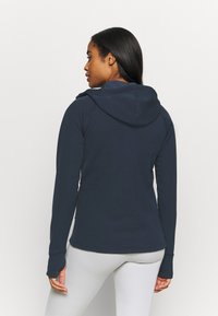 Sweatshirt azul marinho com um design ajustado, mangas longas e capuz com cordão. Apresenta buracos para os polegares nas mangas e uma textura suave.