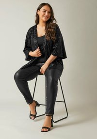 Kimono noir à sequins avec des manches trois-quarts, associé à un top noir et un pantalon en cuir noir ajusté, porté avec des talons à lanières.