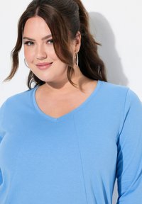 Himmelblaues V-Ausschnitt-Oberteil aus weichem Stoff mit subtiler Textur. Verfügt über eine leicht ausgestellte Silhouette und genähte Details entlang der Nähte.