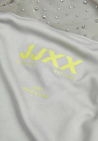 Jasnoszara tkanina z rozsianymi metalicznymi ćwiekami i neonowożółtym logo z napisem "JJXX STUDIOS EST. 2020", wskazującym rozmiar S oraz pochodzenie.