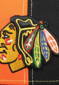 Embroidered patch featuring a stylized head, primarily yellow with red accents, on a black background with green and orange feather details.