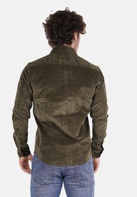 Camicia verde in velluto a coste con maniche lunghe, polsini con bottoni e un dettaglio a yoke angolato; abbinata a jeans blu, vista da dietro.