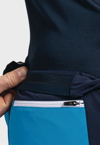 Blå sportshorts med en vit dragkedjeficka och en mörk midja. Hand som greppar midjan, vilket visar den elastiska texturen och designen.