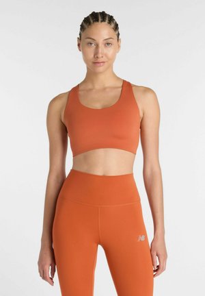 Kvinde med flettet hår iført orange sports-bh og høj taljede leggings, der står foran en ensfarvet hvid baggrund.