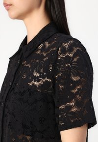 Chemisier en dentelle noire avec un motif floral, des manches courtes et un col pointu. Le tissu est transparent avec des détails délicats tout au long.