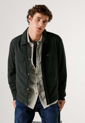 Mladić s kovrčavom kosom nosi tamnu jaknu preko karirane košulje i tamne traperice, blago se naslanjajući prema naprijed na bijeloj pozadini.