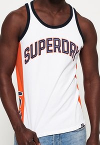 Camiseta sin mangas blanca con panel lateral naranja, con la palabra "SUPERDRY" en letras gruesas. Hecha de tela suave con un escote negro acanalado.