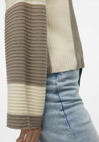 Pull tricoté à rayures beige et taupe. Caractérisé par une texture côtelée et une coupe ajustée avec des manches larges, associé à un jean bleu clair.