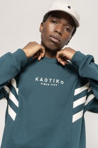 Teal sweatshirts med hvide stribede accenter på ærmerne, med "KAOTIKO SINCE 1999" logo i hvid. Blød tekstur, rund hals design.