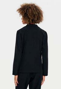 Černý blazer s dlouhými rukávy, výstřihem do V a hladkým povrchem. Obsahuje centrální šev na zadní straně a přiléhavý střih.
