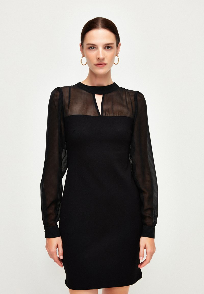 Schwarzes Kleid mit taillierter Silhouette, durchsichtigen karierter langen Ärmeln und einem Ausschnitt mit Schlüsselloch-Detail. Weicher Stoff mit minimalen Akzenten.