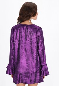 Lila Metallic-Kleid mit langen Ärmeln, gerafften Manschetten und einem ausgestellten Saum. Glatte Textur mit lichtreflektierendem Glanz.