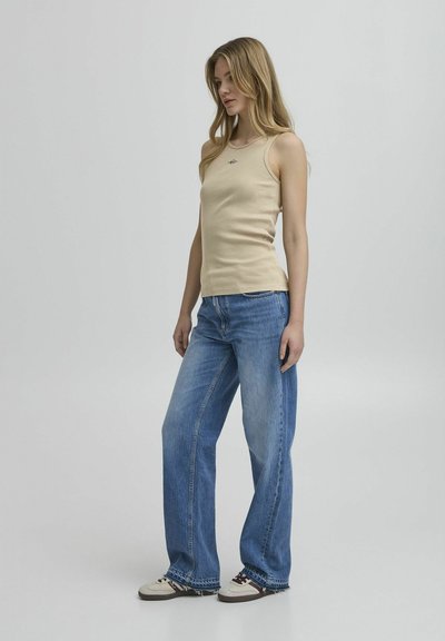 Top de tirantes de canalé beige combinado con vaqueros de pierna ancha en azul claro, con un logo sutil en el pecho y zapatillas deportivas con un diseño en crema y burdeos.