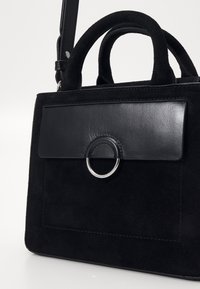 Schwarze Tasche aus Wildleder mit einem glatten Lederdeckel. Verfügt über ein rundes silbernes Designelement und zwei Griffe. Minimalistisches rechteckiges Design.