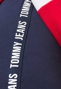 Marinblå sweatshirt med röda och vita detaljer. Har en tygetikett med "TOMMY JEANS" i vit text längs sidan. Mjuk textur.