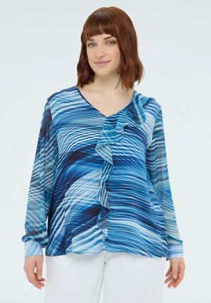 Žena s hnědočervenými vlasy po ramena, oblečená v modré blůze s vlnkovým vzorem a volánkem vpředu, a v bílých kalhotách na jednobarevném pozadí.