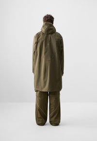 Personne debout de dos, portant une longue veste à capuche vert olive et un pantalon large sur un fond clair uni.
