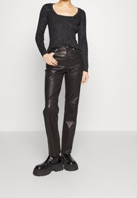 Nainen, joka on pukeutunut mustaan teksturoituun pitkähihastoreeseen, korkeavyötäröisiin mustiin nahkahousuihin ja mustiin paksupohjaisiin bootsiin, seisoo käsi taskussa.