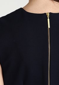 Marinblå klänning med rund halsringning, korta ärmar och en synlig guldlåsning baktill, med ett slätt, strukturerat tyg.