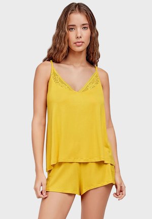 Young woman with wavy hair wearing a yellow sleeveless top with lace trim and matching shorts against a plain background.